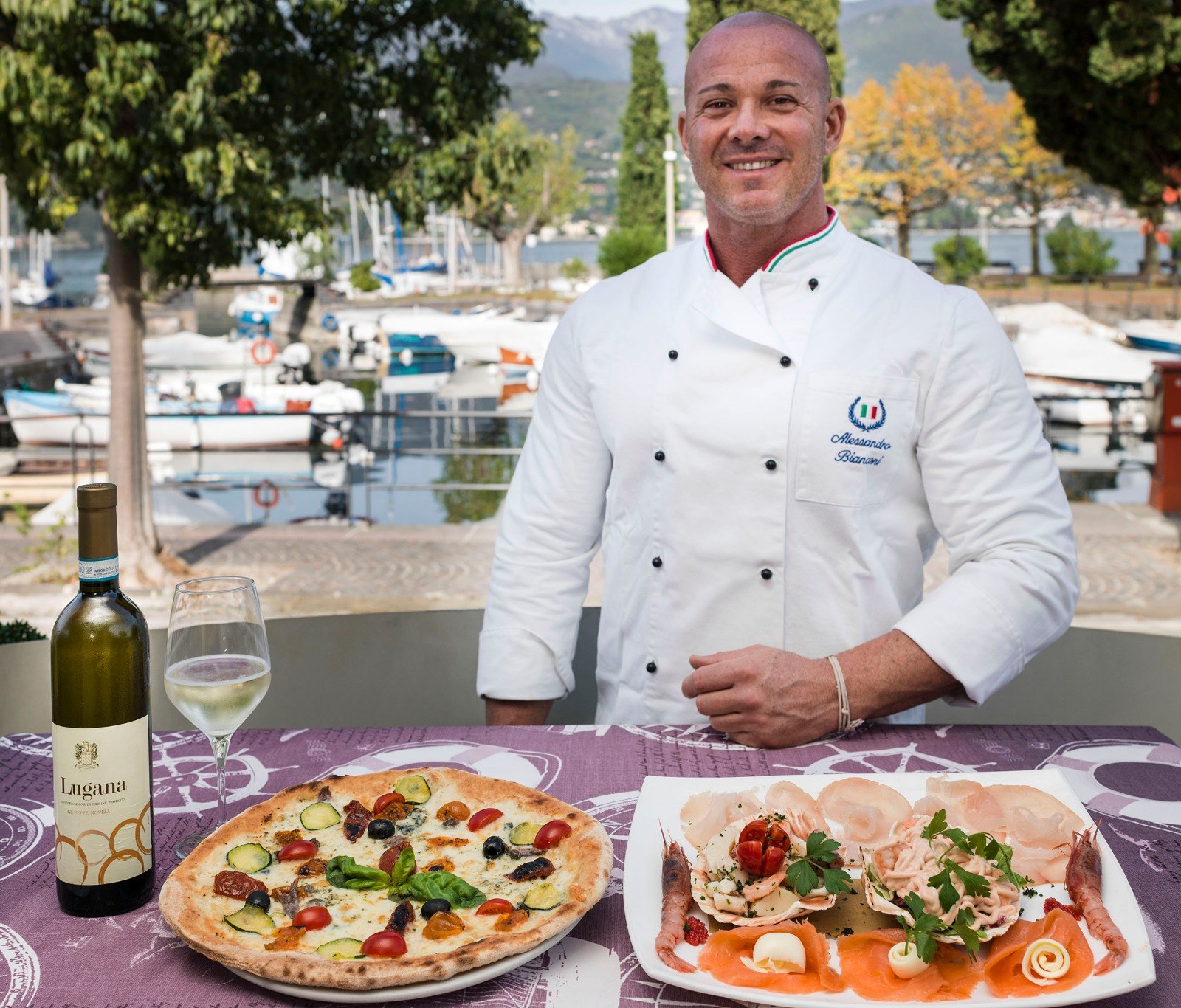 Der Koch an einem Tisch am See, mit einer Pizza, einer Vorspeise und einer Flasche Wein.
