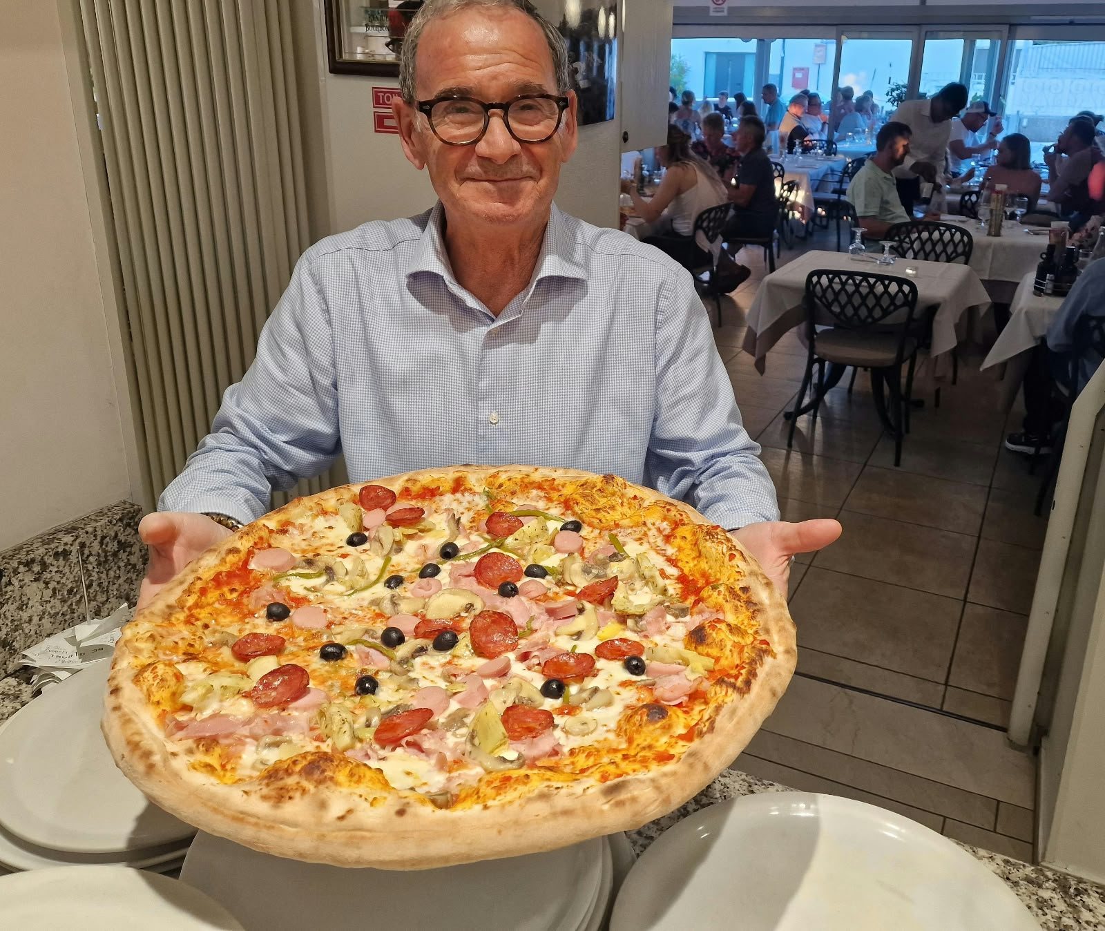 Eine große Pizza im Speisesaal, dahinter die besetzten Tische.