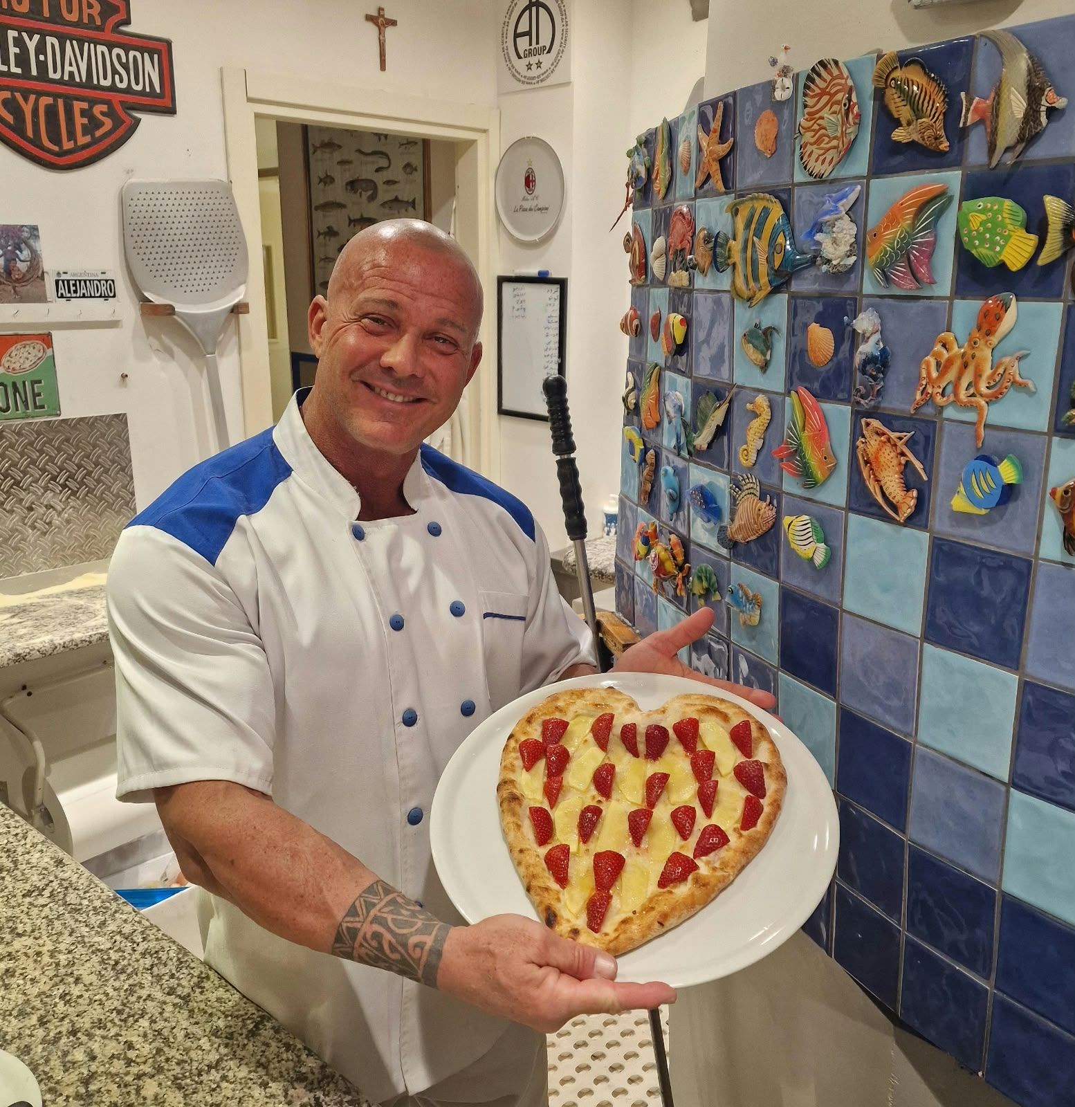 Eine herzförmige Pizza mit Kirschtomaten, vor den blauen Küchenfliesen.
