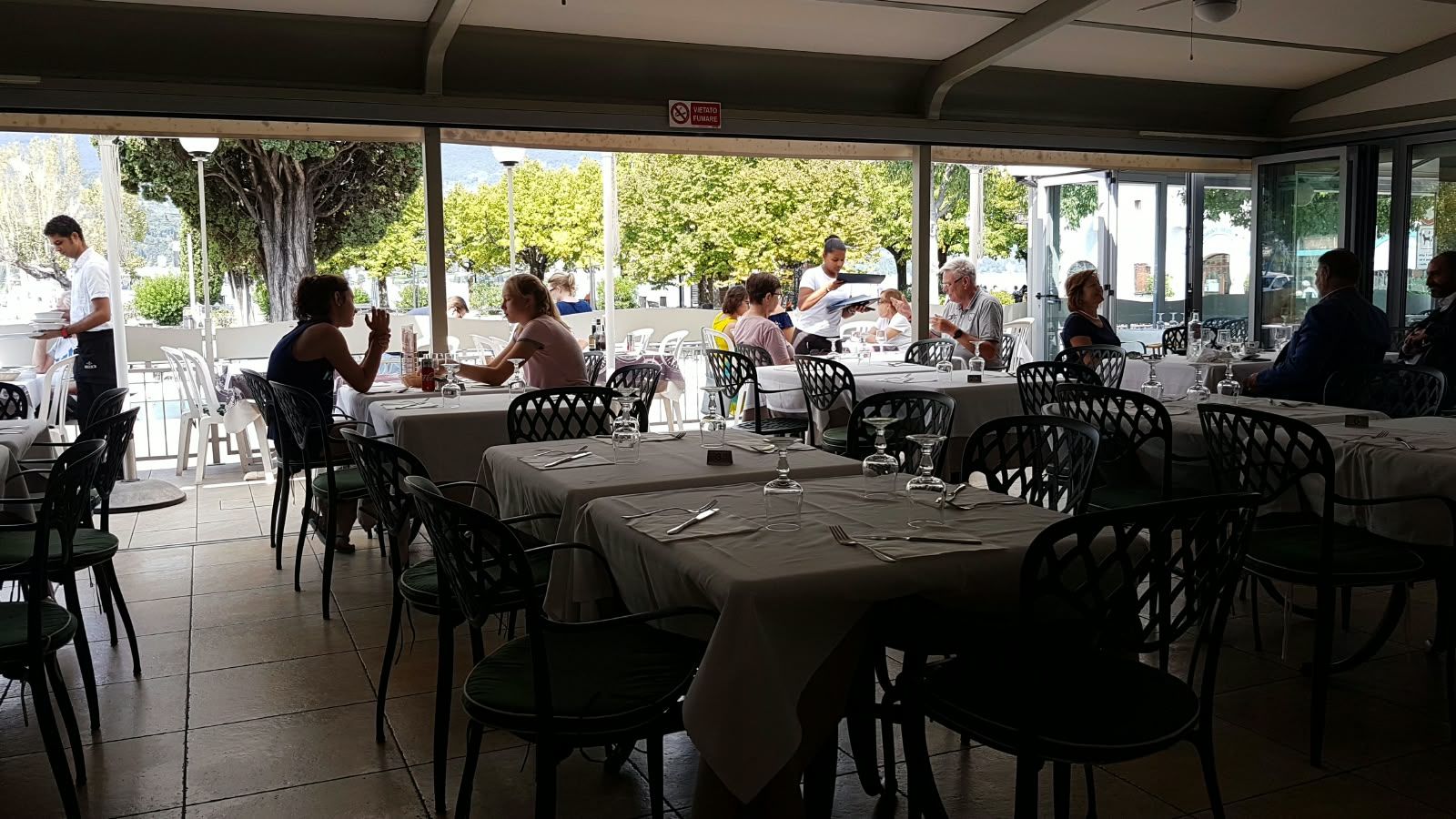 The dining room open to the terrace: laid tables, guests at lunch and the harbour beyond the windows.